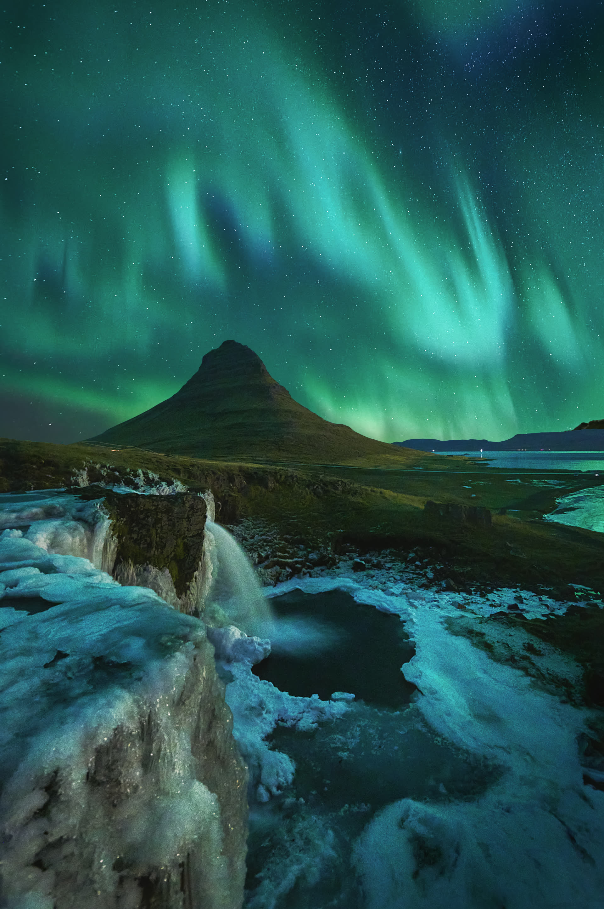 Aurora boreal sobre el monte Kirkjufell desde Kirkjufellfoss, Snæfellsnes, Islandia — composición de dos exposiciones con coche iluminando primer plano